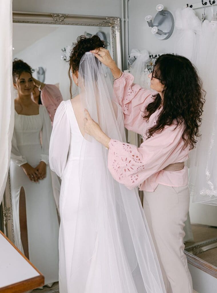 Bride standing at a bridal fitting while a seamstress gently adjusts her veil, reflected in the mirror.