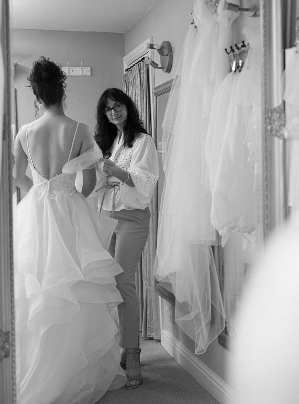 Bride standing at a bridal fitting while a seamstress adjusts the layers of an organza and lace wedding dress, reflected in the studio mirror.